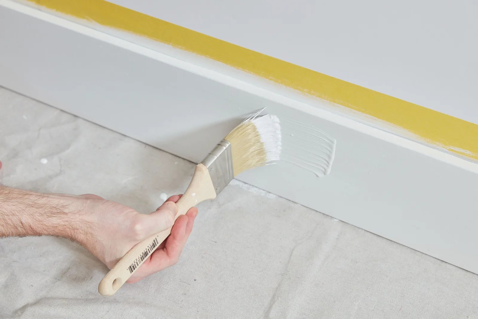 Brush cutting in white paint along a baseboard below a line of painter's tape