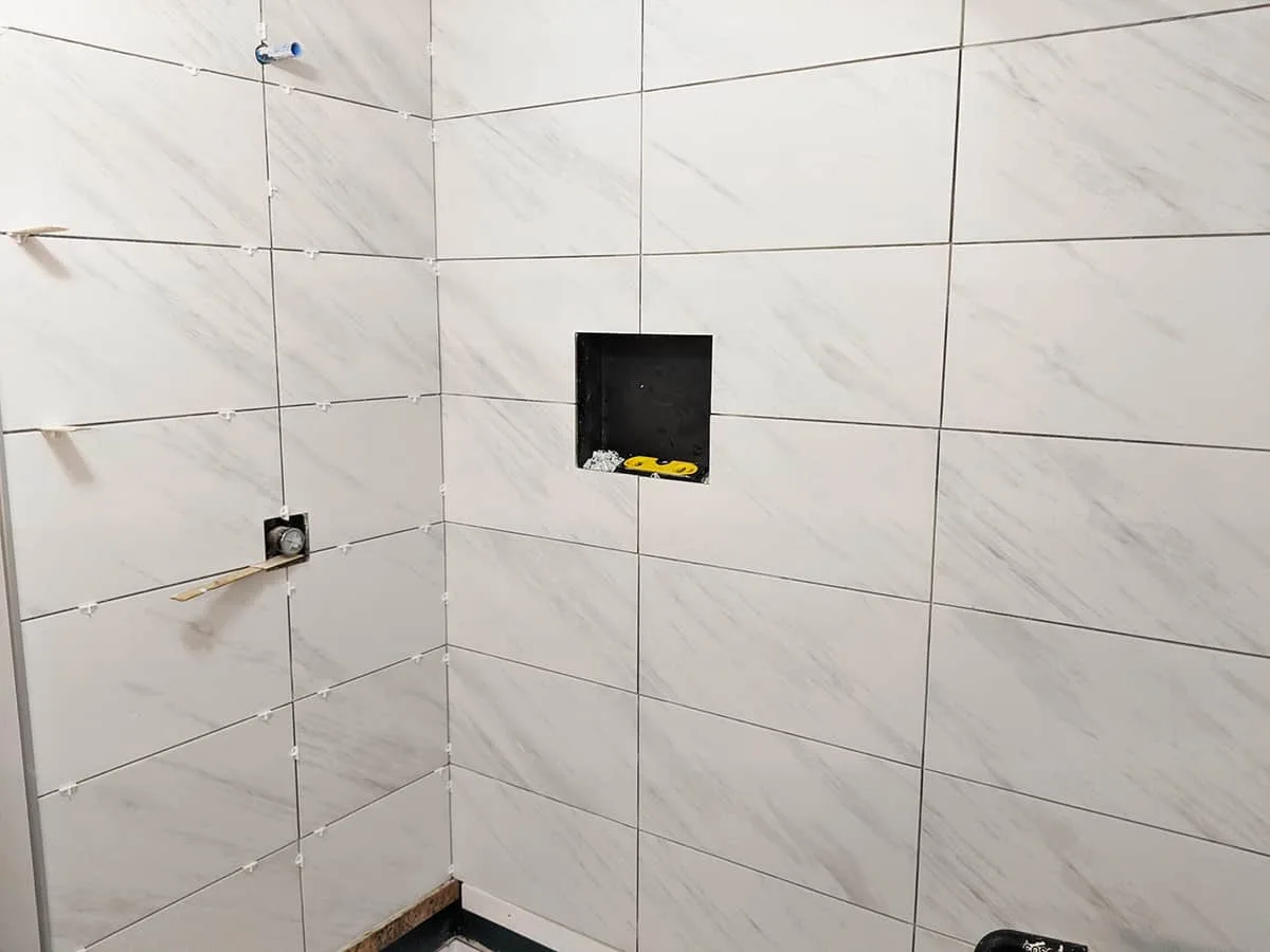 Large-format marble-look wall tile going up in a shower with spacers, a framed niche and the valve rough-in exposed
